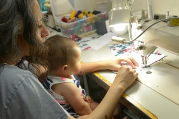 Sewist working on a garment piece on a sewing machine while holding baby